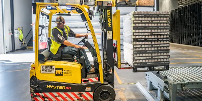 Gabelstaplerfahrer verl&auml;dt Getr&auml;nkestapel auf F&ouml;rderband in moderner Industriehalle. Effiziente Lagerlogistik.